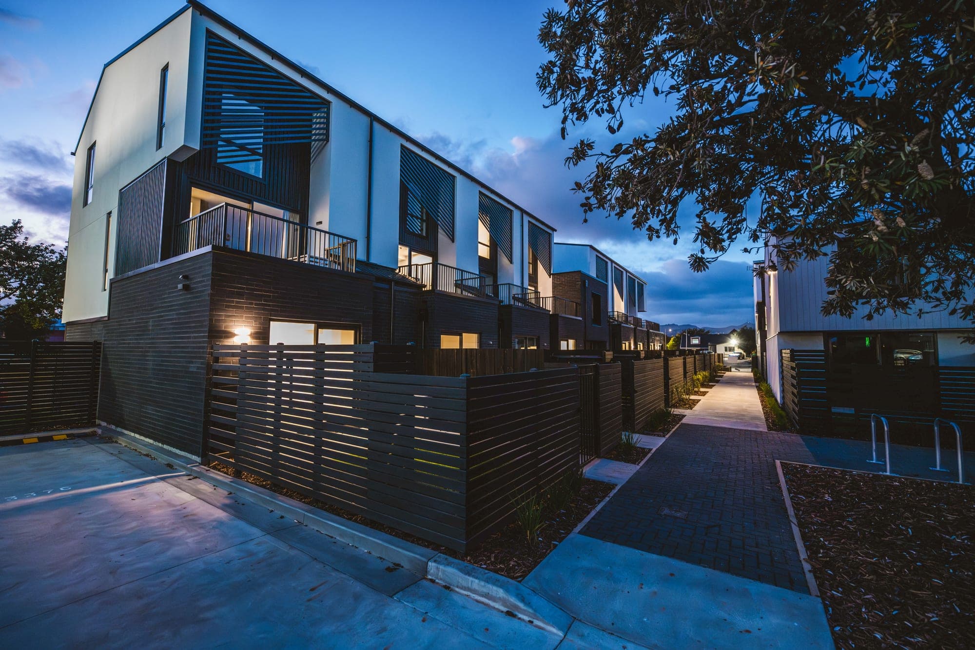 The street frontage at dusk — concrete aprons and paver footpath running the length of the development.