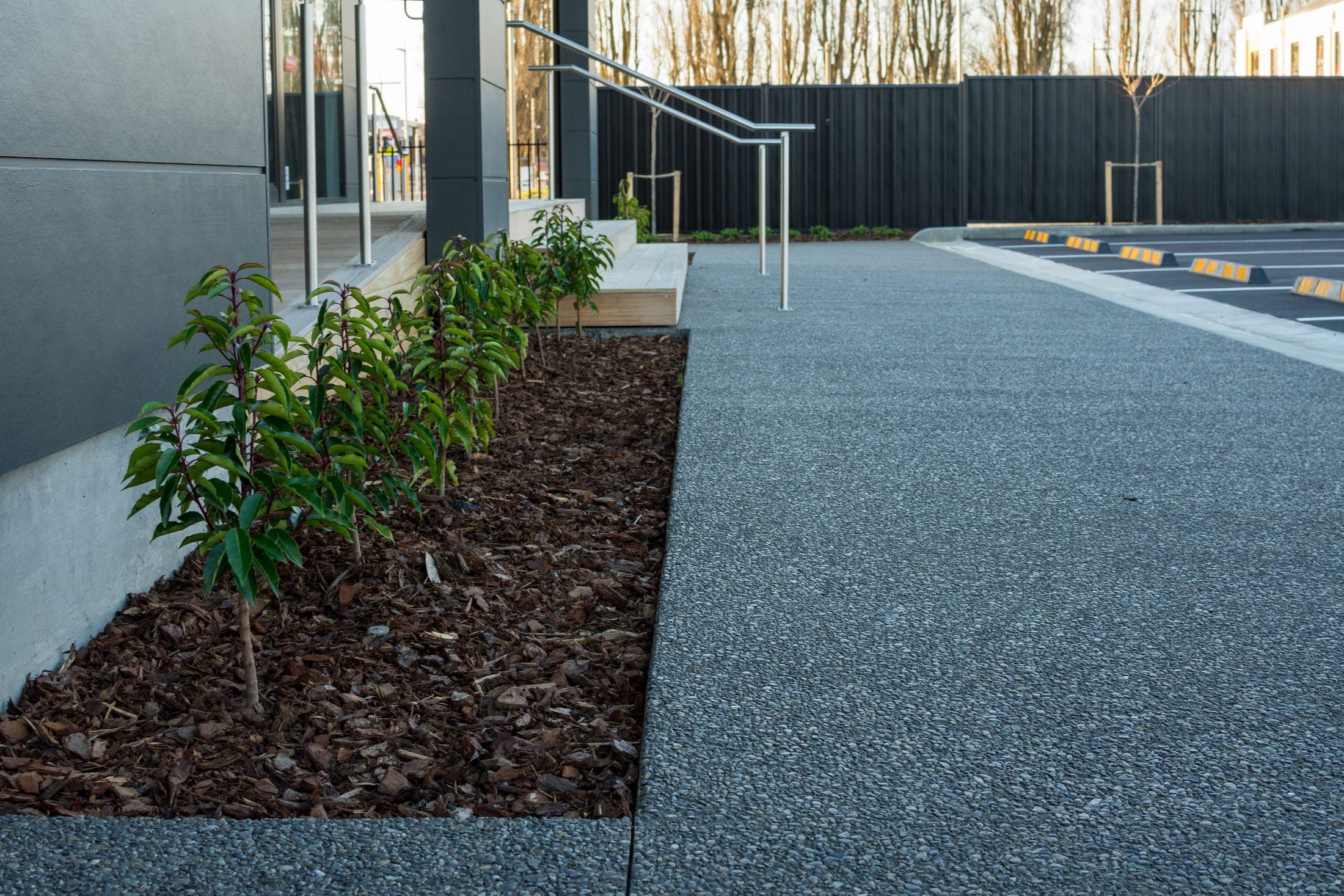Exposed-aggregate footpath and planter surrounds running along the building to the accessible entry ramp.
